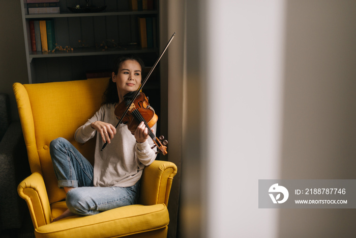 Fiddler plays tender music on a violin in her home near the window. Attractive young woman musician plays the violin sitting on soft chair.