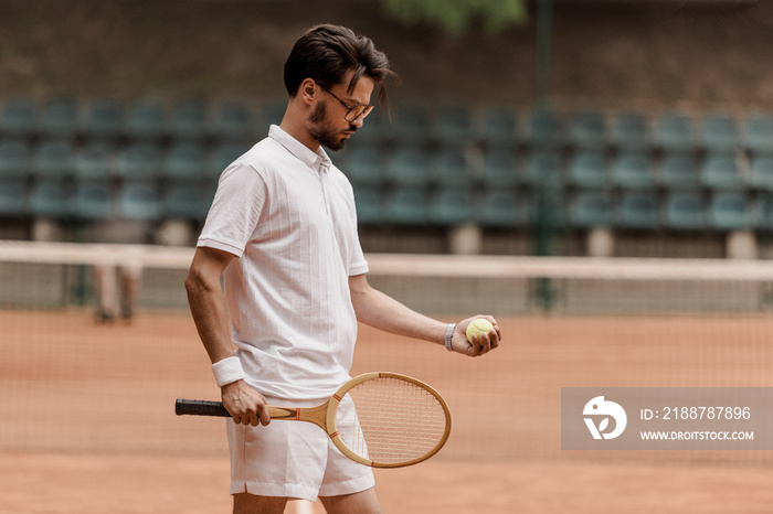 handsome retro styled tennis player standing with tennis ball and racket at court