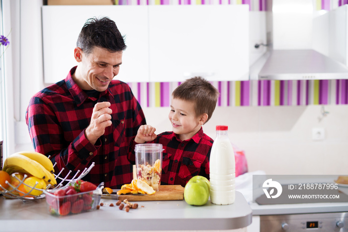 Young happy father and son are putting diverse cut fruit into the bowl preparing it for blending with milk on the kitchen counter.