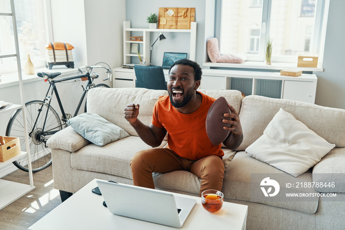 Carefree young African man cheering and smiling while watching sport match at home