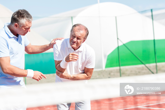 Mature man helping senior friend with elbow injury while playing tennis on sunny day