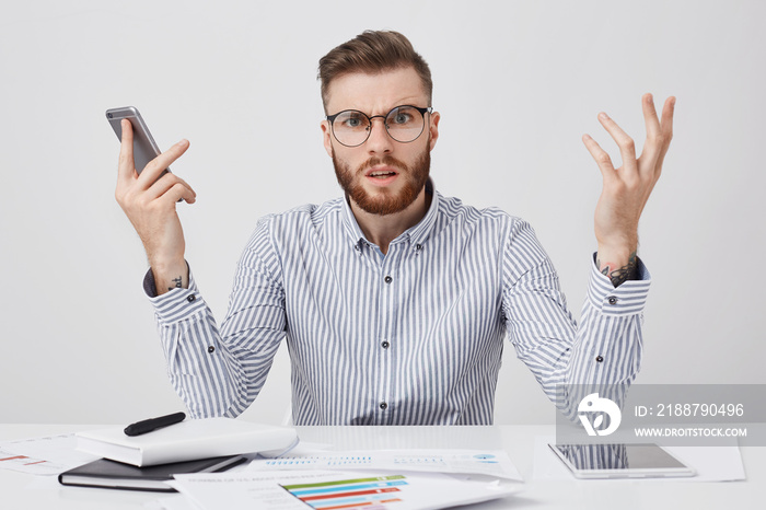 Puzzled bearded man with indignant look can`t understand why there is no answer from colleague, gestures in furious manner, keeps mobile phone in hand, surrounded with documents and tablet computer