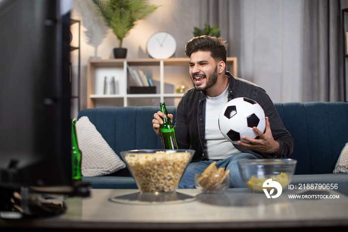 Excited indian man with chips and beer sitting on cozy sofa and watching sport TV. Young bearded guy supporting favorite football team during world cup.