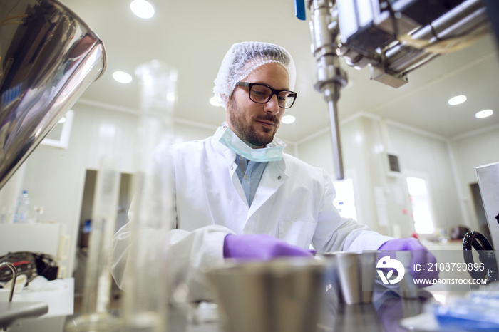 Close up of serious focused professional scientist bearded young man sitting and working at laboratory machine with metal flasks and bottles in front.