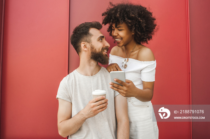 happy young multiethnic couple with smartphone and coffee to go smiling each other on street