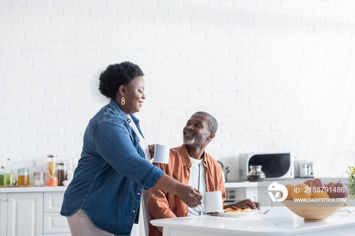 happy african american woman holding cups of coffee near smiling husband during breakfast.