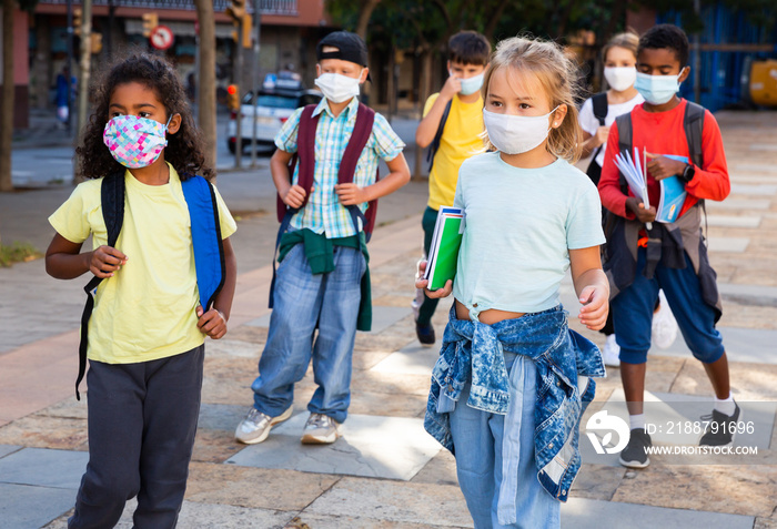 Group of schoolchildren with backpacks and exercise books wearing face masks going together to open air lesson