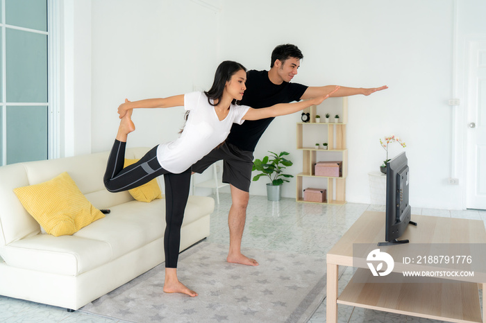 Young Asian couple doing yoga practice together and looking TV at home, man and woman working out together standing in living room, fit pair performing fitness exercise with partner.