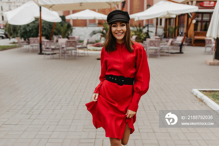 Portrait of smiling girl with dark eyes and brown hair. Lady in red silk dress walks easily in Parisian street