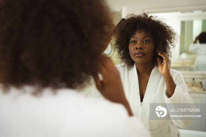 African american woman in bathroom wearing bathrobe, looking in mirror