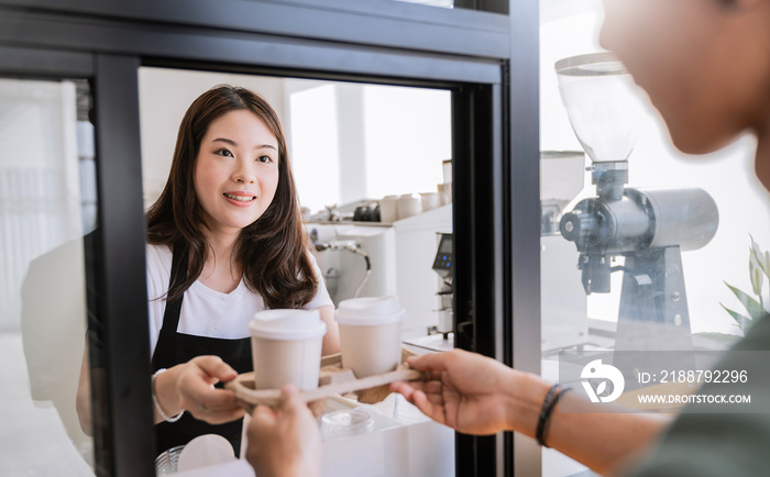 Young asia woman hands gives paper coffee cup on slide glass window. Take away delivery concept. Asian woman barista. Cropped shot of waitress and buyer hold coffee, eco recycle environment friendly.