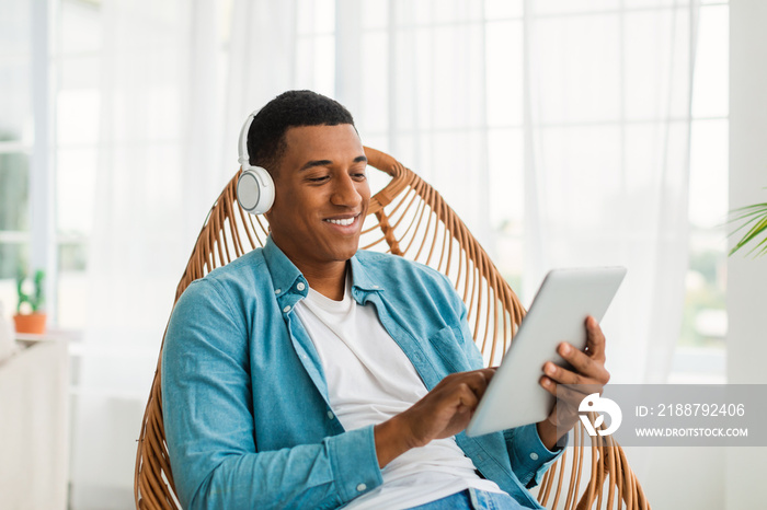 Glad happy young black male in casual and wireless headphones chatting in social networks on tablet