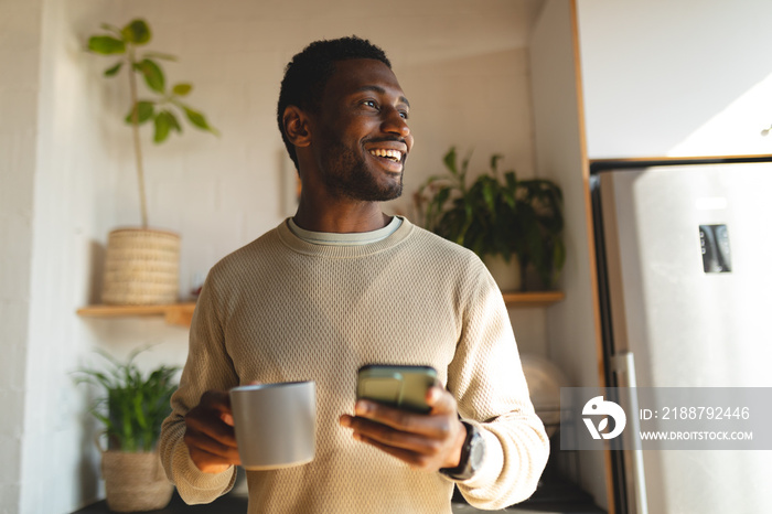 Happy african american man using smartphone and drinking coffee in kitchen