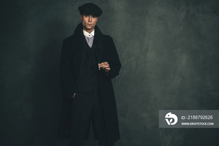 Macho retro 1920s english gangster wearing black coat and flat cap holding cigarette.