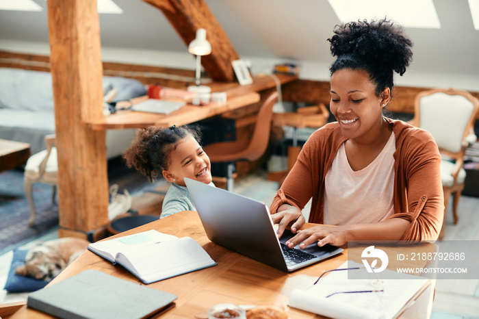 Happy black single mother works on laptop while being with her daughter at home.