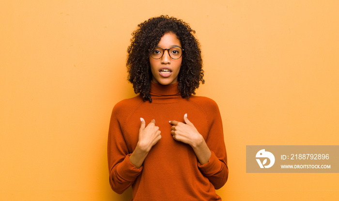 young pretty black woman pointing to self with a confused and quizzical look, shocked and surprised to be chosen against orange wall