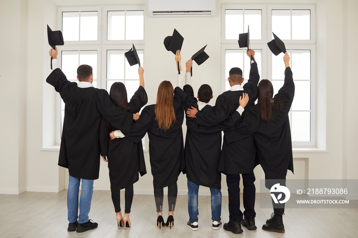 Rear view group of multiracial students in graduation gowns stand in a classroom with their backs to the camera with raised academic mortar boards. Concept of education, graduation and alumni.