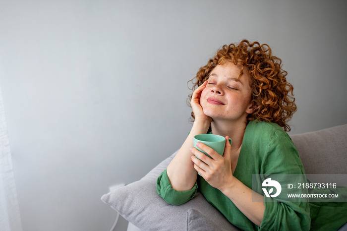 Pretty redhead woman savoring her mug of hot coffee as she sits on a sofa with eyes closed in contentment. Cup of tea or coffee. Woman drinking hot beverage and enjoying morning