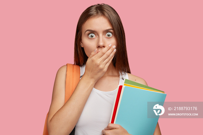 Horizontal shot of attractive shocked young woman wuth bugged eyes, covers mouth and stares with fear, finds out she failed exam, carries rucksack, holds textbooks, dressed in casual t shirt