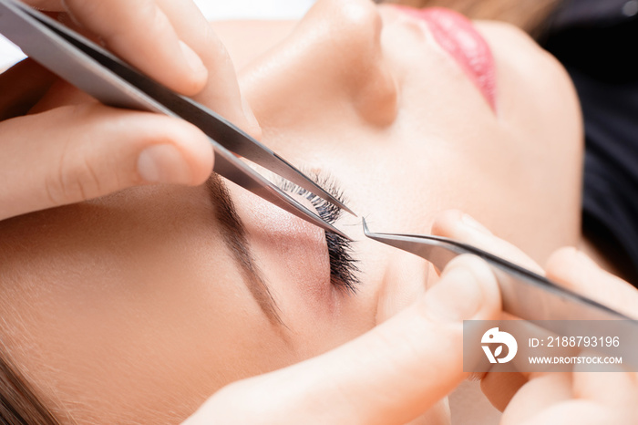 Eyelash extension procedure. Master tweezers sets fake lashes on beautiful woman