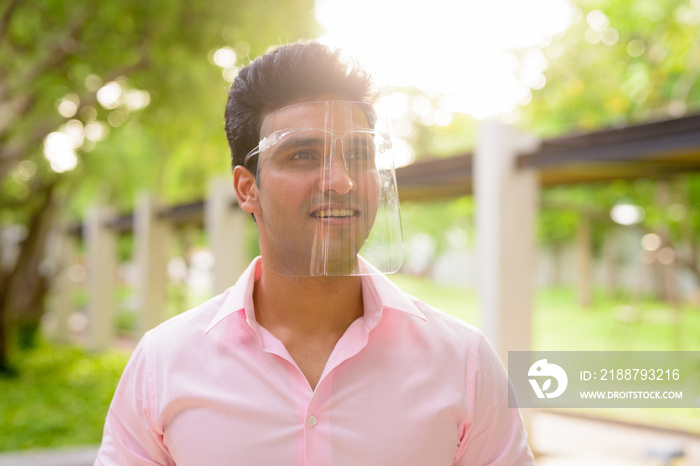 Happy young handsome Indian businessman with face shield thinking at the park