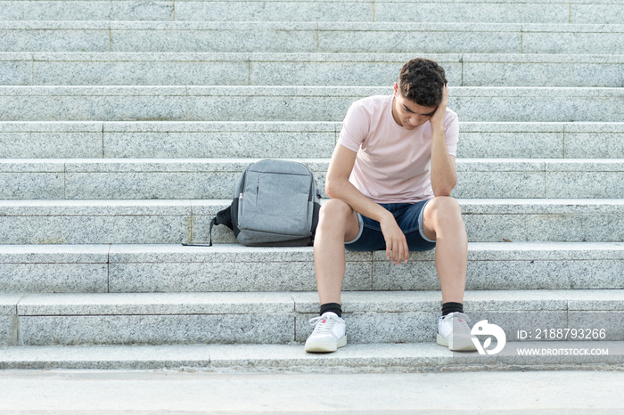 Sad and frustrated hispanic teenager sitting on stairs and holding his head. Anxiety and depression in adolescence concept.