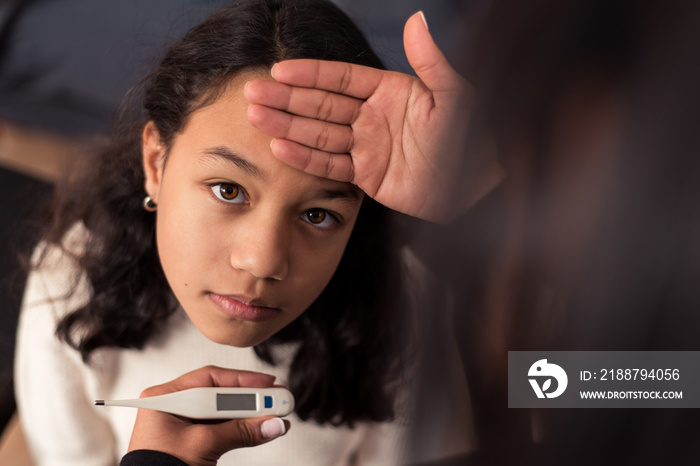 Girl looking at camera while her mother check the temperature with a thermometer at home.