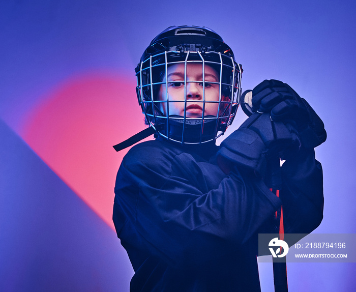 Young blonde fit boy, ice hockey player, posing in a bright neon studio for a photoshoot, wearing an ice-skating uniform while wearing his helmet, holding a hockey stick in his arms and looking