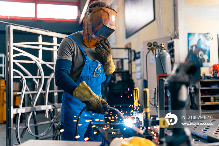 A worker is using a welding machine in his hand and working on a metal rod, he is wearing his visor for protection