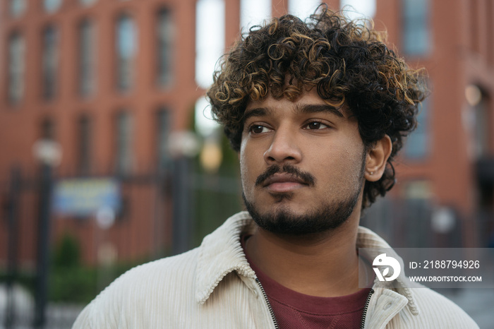 Closeup portrait of pensive Indian man with curly hair wearing stylish jacket looking away on the street