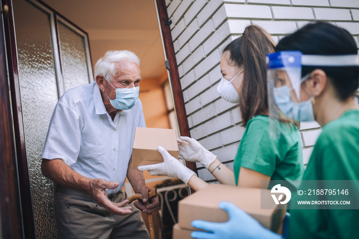 Young female volunteers in mask gives an elderly man boxes with food near his house. Quarantined, isolated. Coronavirus covid-19. Donation