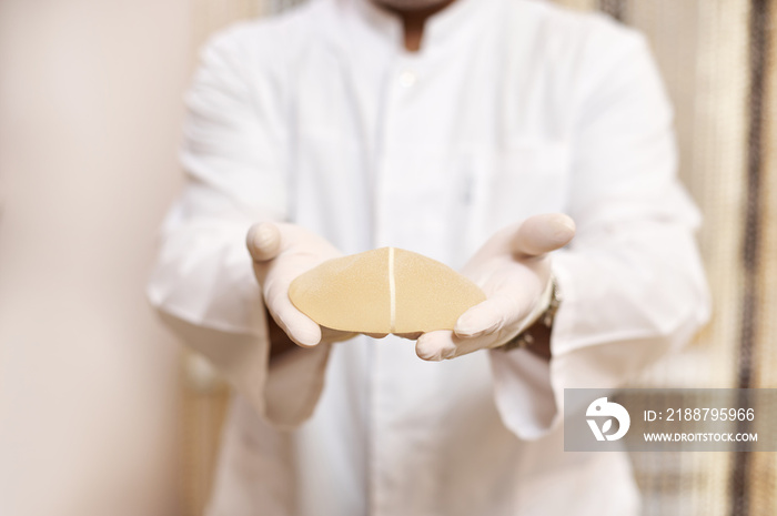 Plastic surgeon holds breast implant and shows it to the camera standing on the background of wall in his cabinet