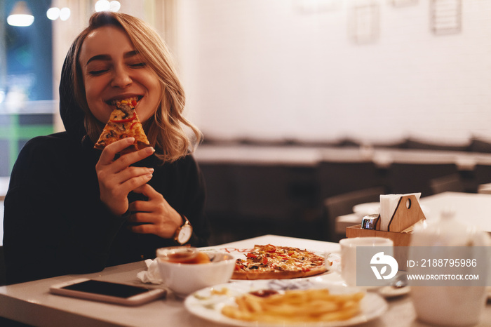 Funny blonde girl in black sweater with hood eating pizza at restaurant.