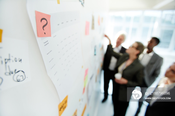 Multi-ethnic team of talented white collar workers gathered together at marker board while having joint project discussion at spacious open plan office, focus on foreground