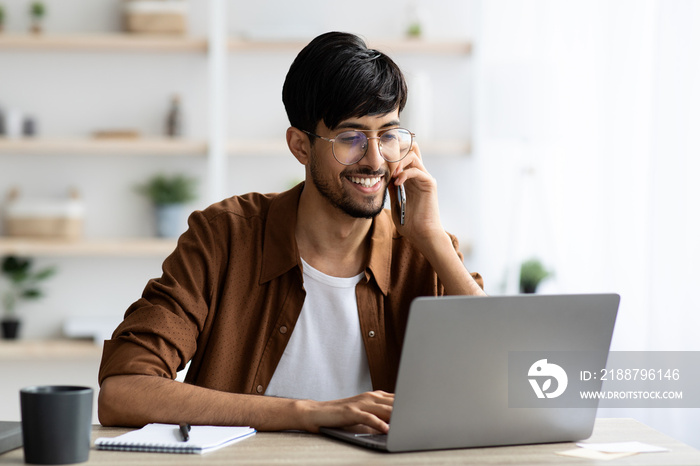 Cheerful indian guy businessman with laptop having phone conversation