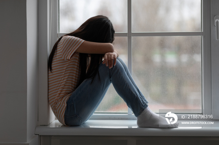 Upset long-haired brunette girl sitting on windowsill and crying