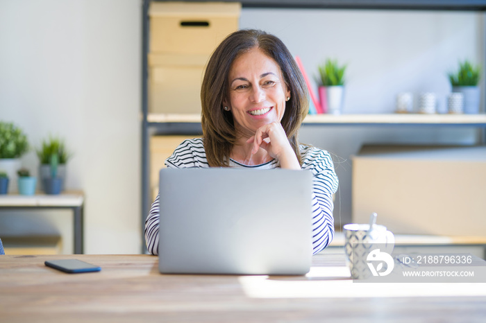 Middle age senior woman sitting at the table at home working using computer laptop looking confident at the camera smiling with crossed arms and hand raised on chin. Thinking positive.