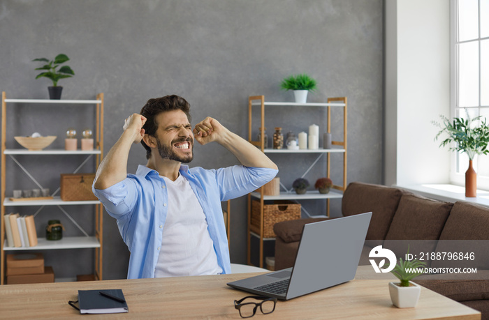 Yes, I did it. Young man sitting at working desk in front of laptop computer, feeling excited, smiling and fist pumping. Emotional student passes exam, gets excellent grade and is happy about success