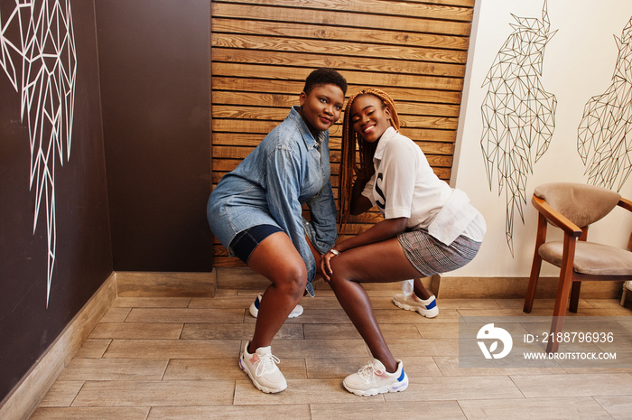 Two african womans in stylish casual clothes posing against wooden wall sat down and dance twerk.