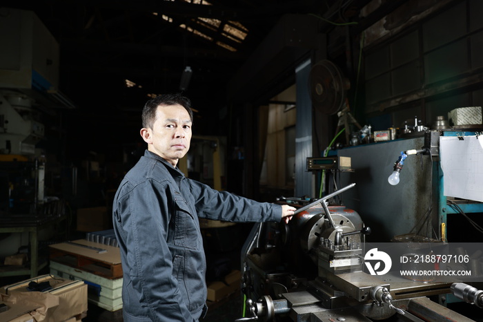 Metalworker in gray work clothes and a lathe in a small to medium-sized small business town factory. Conceptual images of the essence of manufacturing and technical succession.