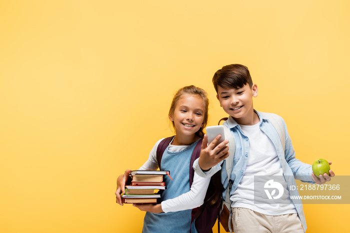 Smiling multiethnic pupils with books and apple using mobile phone isolated on yellow.