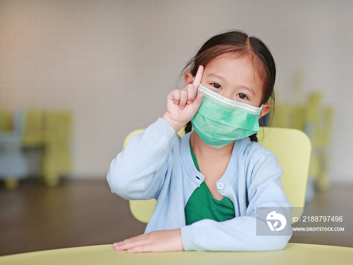 Cute little Asian child girl wearing a protective mask with showing one forefinger sitting on kid chair in children room.