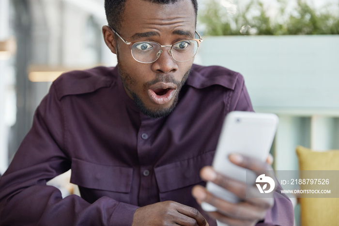 Shocked dark skinned young man in spectacles surprised as gets bill receipt on mobile phone, stressed to read shocking news, has business problems, reacts emotionally, worried about something