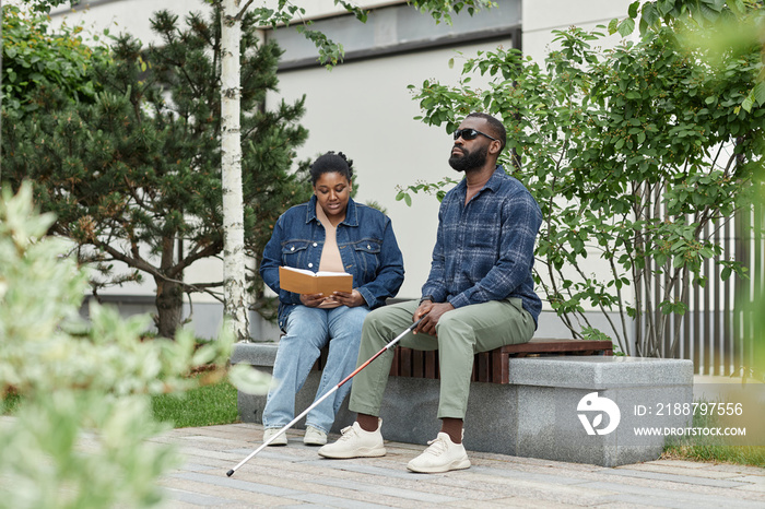 Full length portrait of adult couple with partner with visual disability sitting on bench in park together