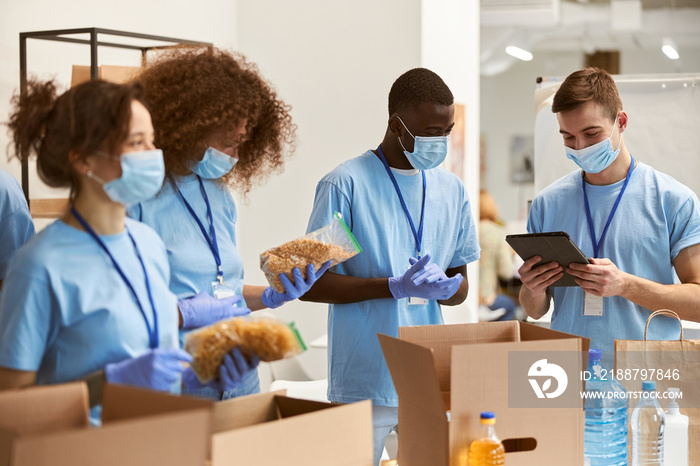 Team of volunteers in protective masks and gloves sorting, packing food in cardboard boxes, working on donation project during pandemic