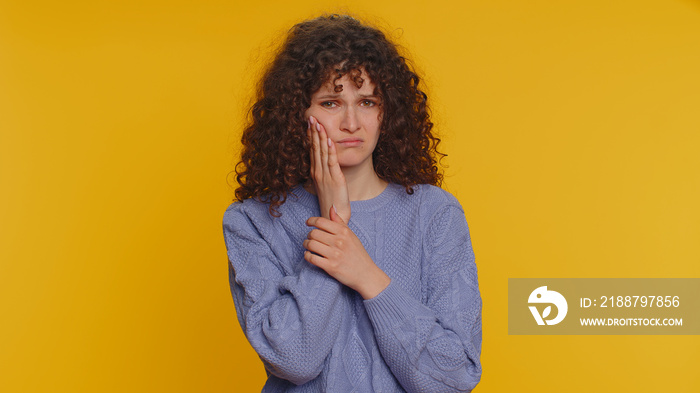 Hipster curly haired woman touching sore cheek suffering from toothache cavities or gingivitis waiting for dentist appointment gums disease. Young girl indoor studio shot isolated on yellow background