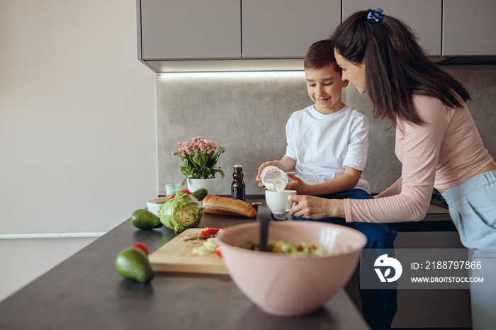 Young beautiful mother showing to small son how to cook and boy pouring milk in cup.