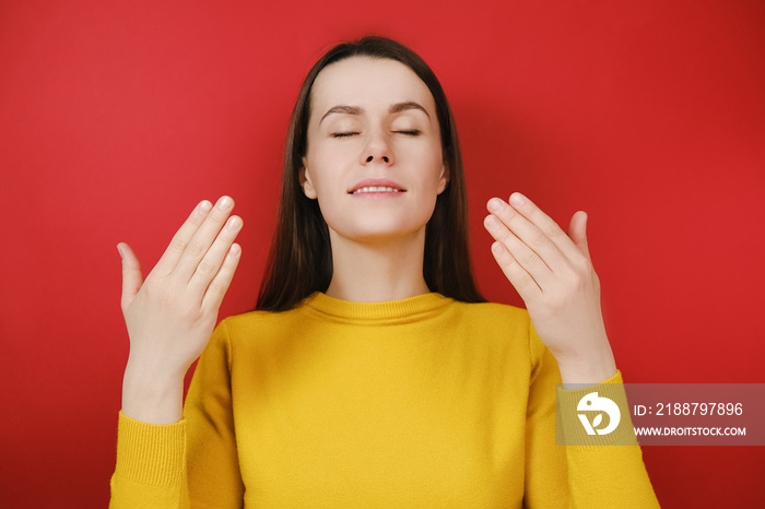 Relaxed young female model enjoying good smell or pleasant fragrance, serene mindful young female taking deep breath feel no stress free inhaling fresh air relaxing isolated on red studio background