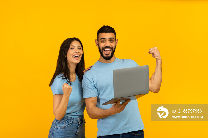 Overjoyed arab couple holding laptop and celebrating success with clenched fists, lucky winners with PC