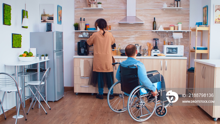 Wife taking groceries bag from paralyzed husband in wheelchair in kitchen after arriving from the market. Paralyzed disabled impairment handicap disability man in wheelchair helping and recovering.